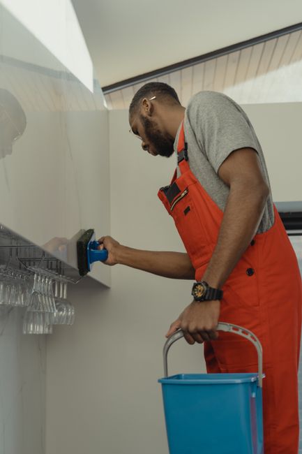 Close-up of a person wearing yellow rubber gloves performing surface cleaning on white ceramic tiles in a kitchen or bathroom. The individual is holding an orange sponge in one hand and a spray bottle