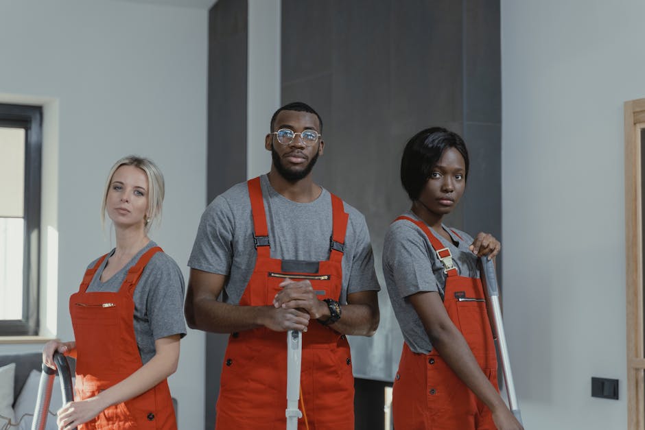 Three professional cleaners from Deep Deep Cleaning standing indoors in a modern, well-lit room. They are wearing matching grey t-shirts underneath bright orange coveralls with black accents, each hol