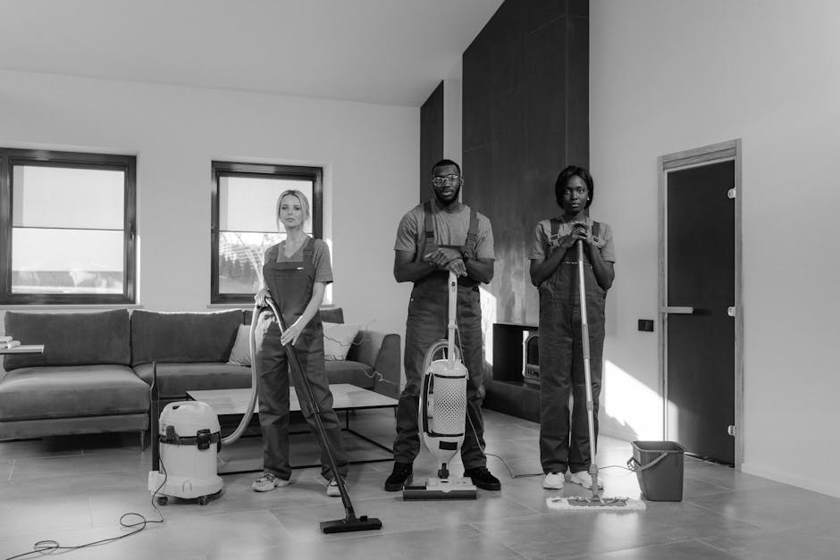 Three professional cleaners from Deep Deep Cleaning standing indoors in a modern, well-lit room. They are wearing matching grey t-shirts underneath bright orange coveralls with black accents, each hol