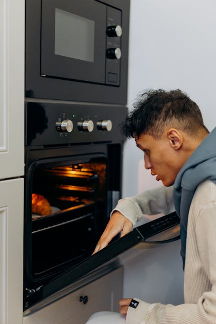 A person with curly hair, wearing a beige long-sleeve top, is opening the oven door of a built-in oven located under a light wooden countertop in a domestic kitchen setting. The oven has a stainless s